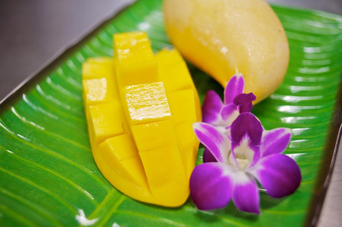 The image shows a plate of sliced mango, a whole mango, and a purple orchid flower, all arranged on a green leaf. The mango is cut into cubes and presented as a dessert.