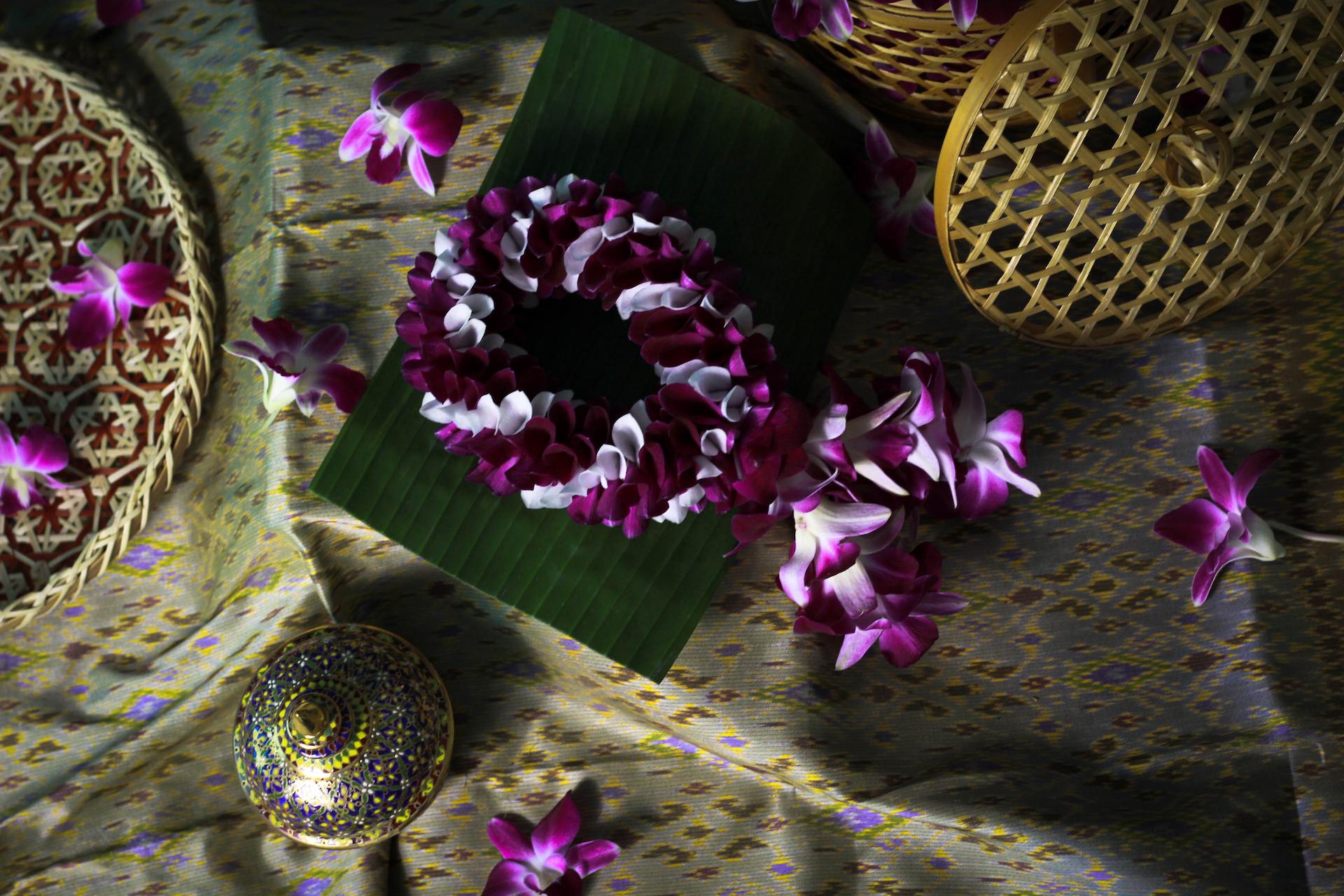 The image showcases a decorative arrangement featuring a floral garland of purple and white orchids, various woven baskets, a decorated container, and scattered orchids on a patterned fabric. The composition has a traditional aesthetic.