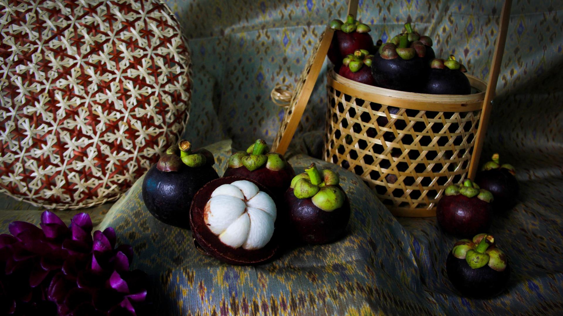 The image displays mangosteen fruit, some whole and some cut open to reveal the white flesh, arranged on a patterned fabric. A woven basket and a decorative floral element are also included in the composition.
