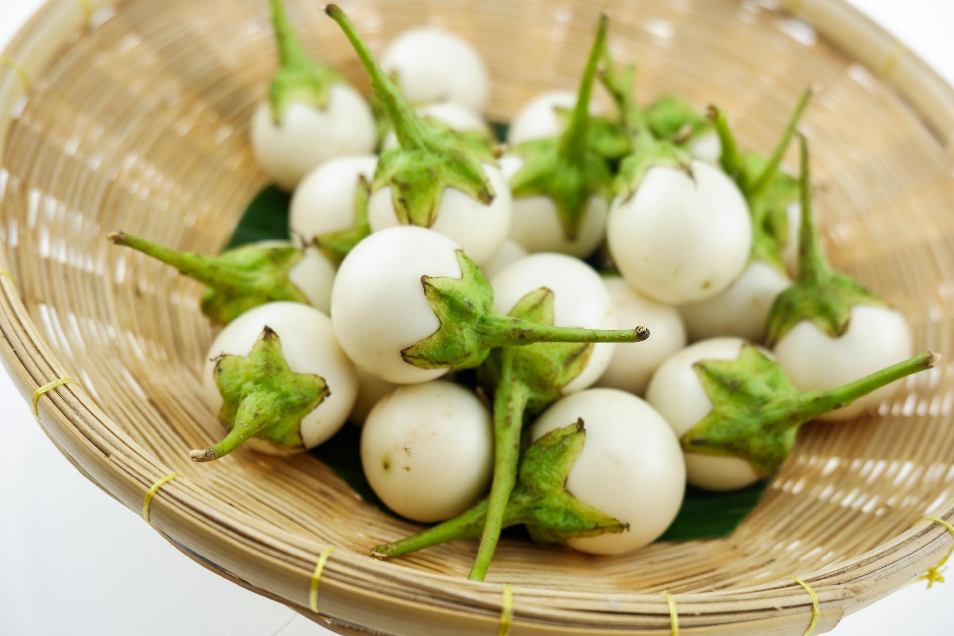 A woven basket is filled with small, white eggplants with green stems. The eggplants are the main focus, with the basket providing a natural background.