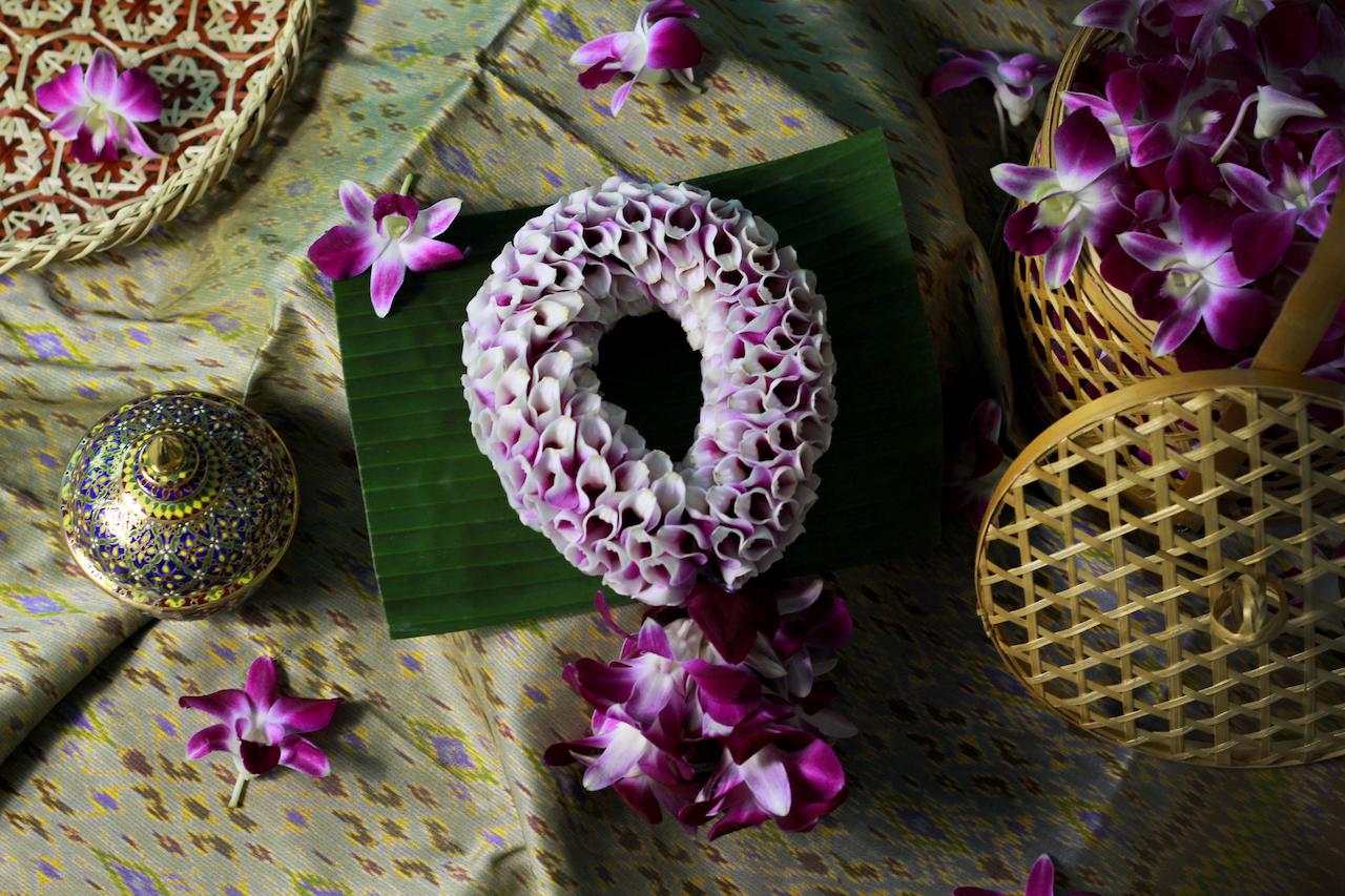 The image showcases a floral arrangement featuring a garland of purple and white flowers. Various decorative elements like baskets, a bowl, and additional flowers are arranged around the garland.